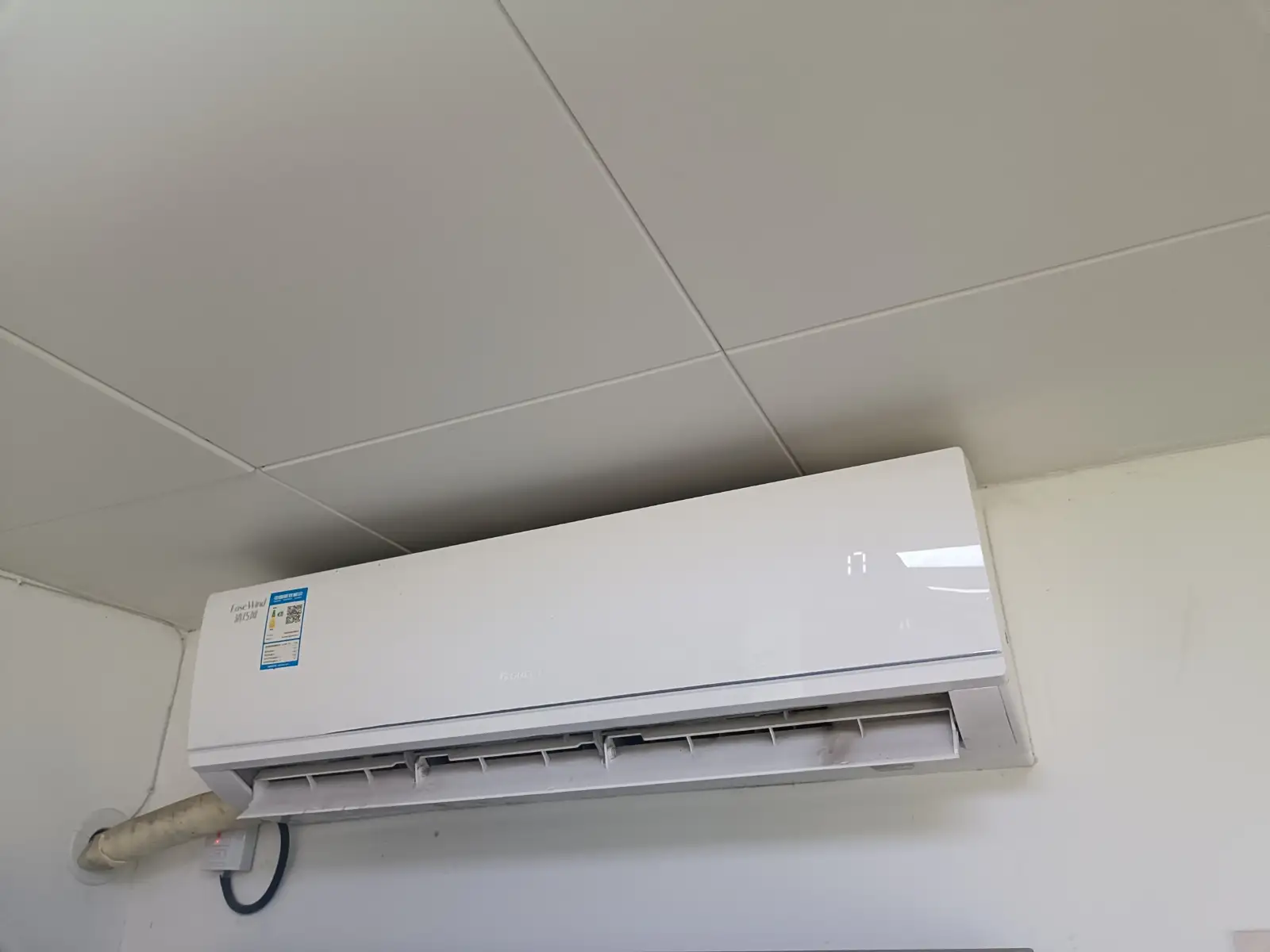 A white air conditioning unit mounted high on a wall, below a tiled ceiling